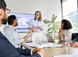 A female leader stands at the front of a conference room, delivering a presentation to a group of colleagues. A female leader stands at the front of a conference room, delivering a presentation to a group of colleagues.