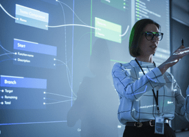 A woman confidently delivering a presentation in front of a projector screen displaying visuals. The room is dark with blue lighting, highlighting her presence as she engages the audience with gestures, showcasing her expertise and poise.