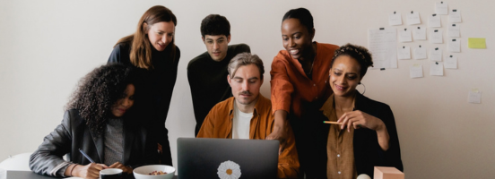 A diverse team of professionals is gathered in an office, collaborating around a laptop. A diverse team of professionals is gathered in an office, collaborating around a laptop.