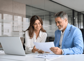 A woman and a man in business attire sitting in a modern office, smiling as they work together.
