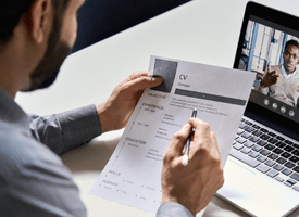 A recruiter sits at a desk, holding and reviewing a printed copy of a candidate's CV during a video interview.