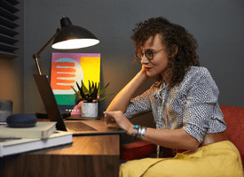 A woman working on a laptop in a cozy home office, seated at a desk with a stylish lamp. She is reclining her head on one hand, appearing focused and relaxed.